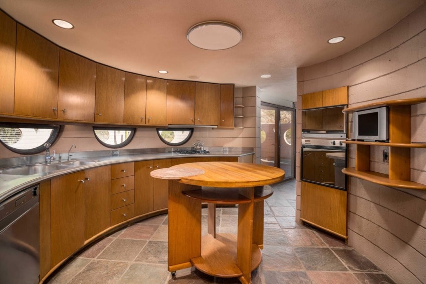Curved kitchen with circular windows, wooden cabinets, round island, built-in microwave, and stone tile floor.