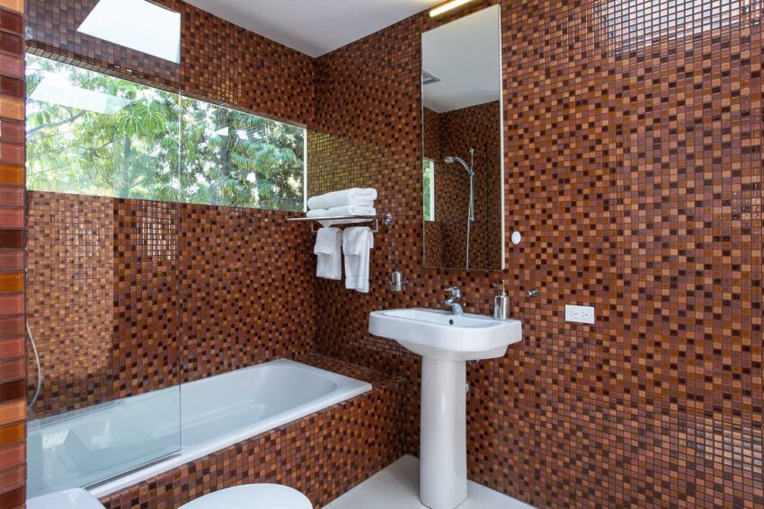 modern bathroom with orange mosaic tile walls, glass shower, bathtub, and white pedestal sink with towels on a wall shelf