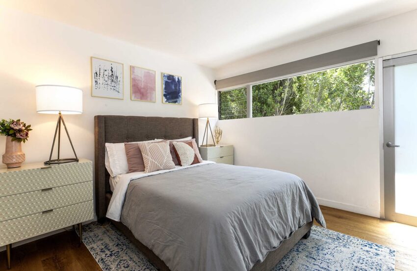 Modern bedroom with a cozy bed, stylish nightstands, soft lighting, artwork on walls, and a window with a view of greenery.