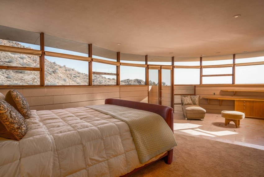 Spacious bedroom with panoramic windows offering a mountain view, featuring a bed, armchair, ottoman, and wooden furniture.
