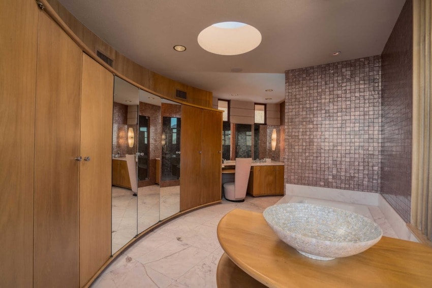 Modern bathroom with wooden curved cabinetry, large mirrors, and a decorative stone bowl on a round wooden table.