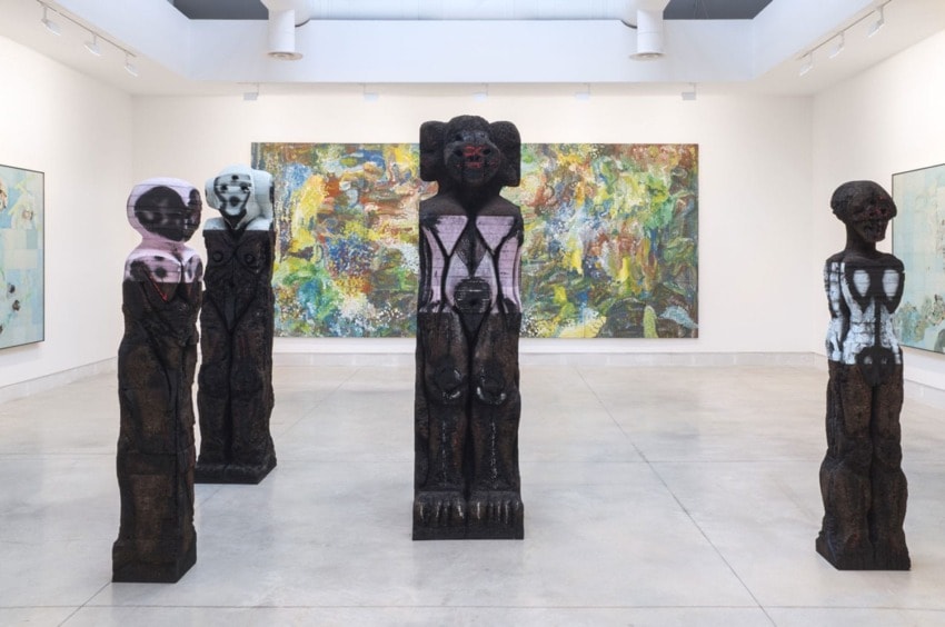 Art gallery with colorful abstract paintings and four tall wooden sculptures standing on a light grey floor.