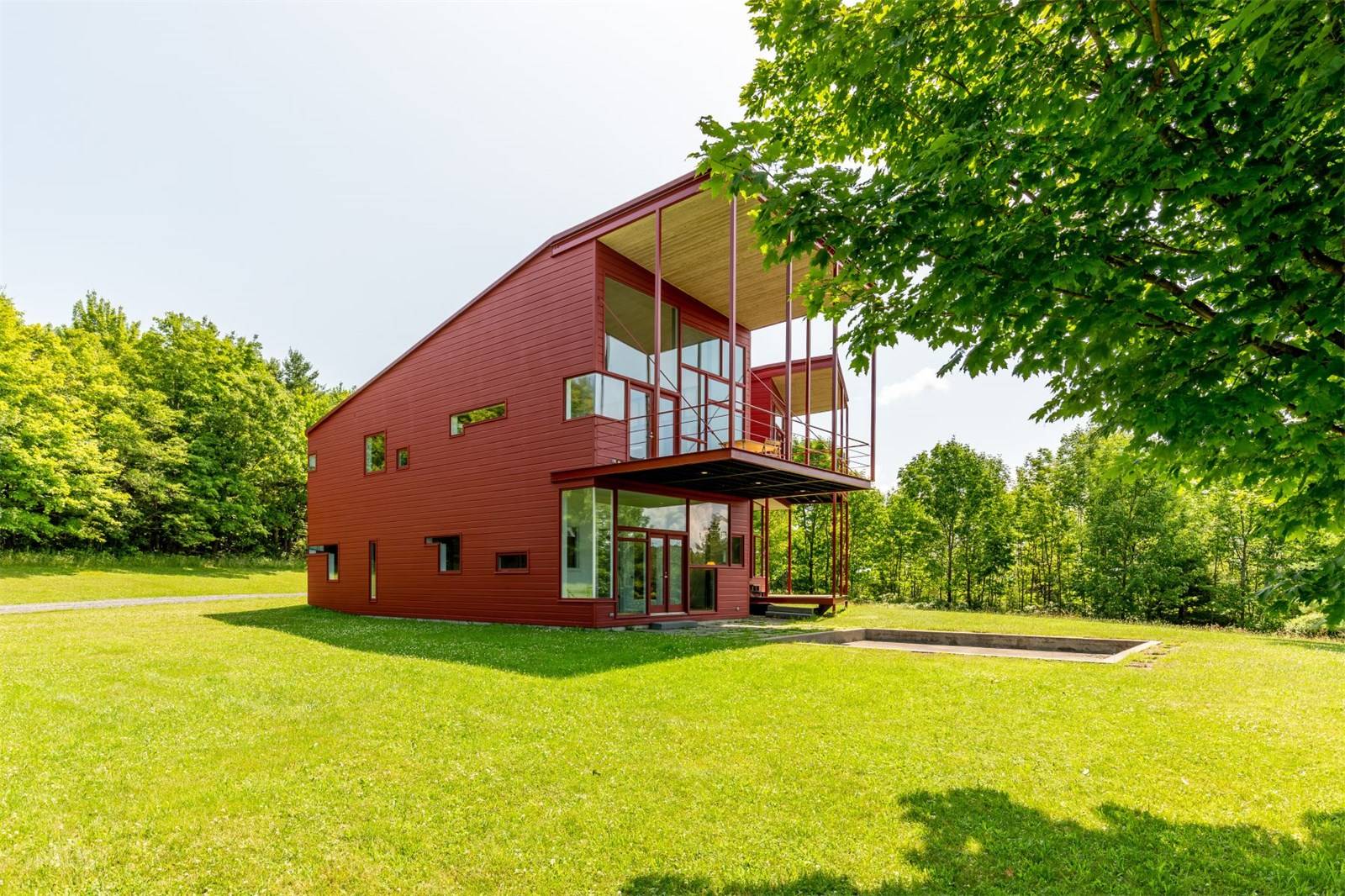 Modern red house with large windows and balconies surrounded by green grass and trees under a clear sky.