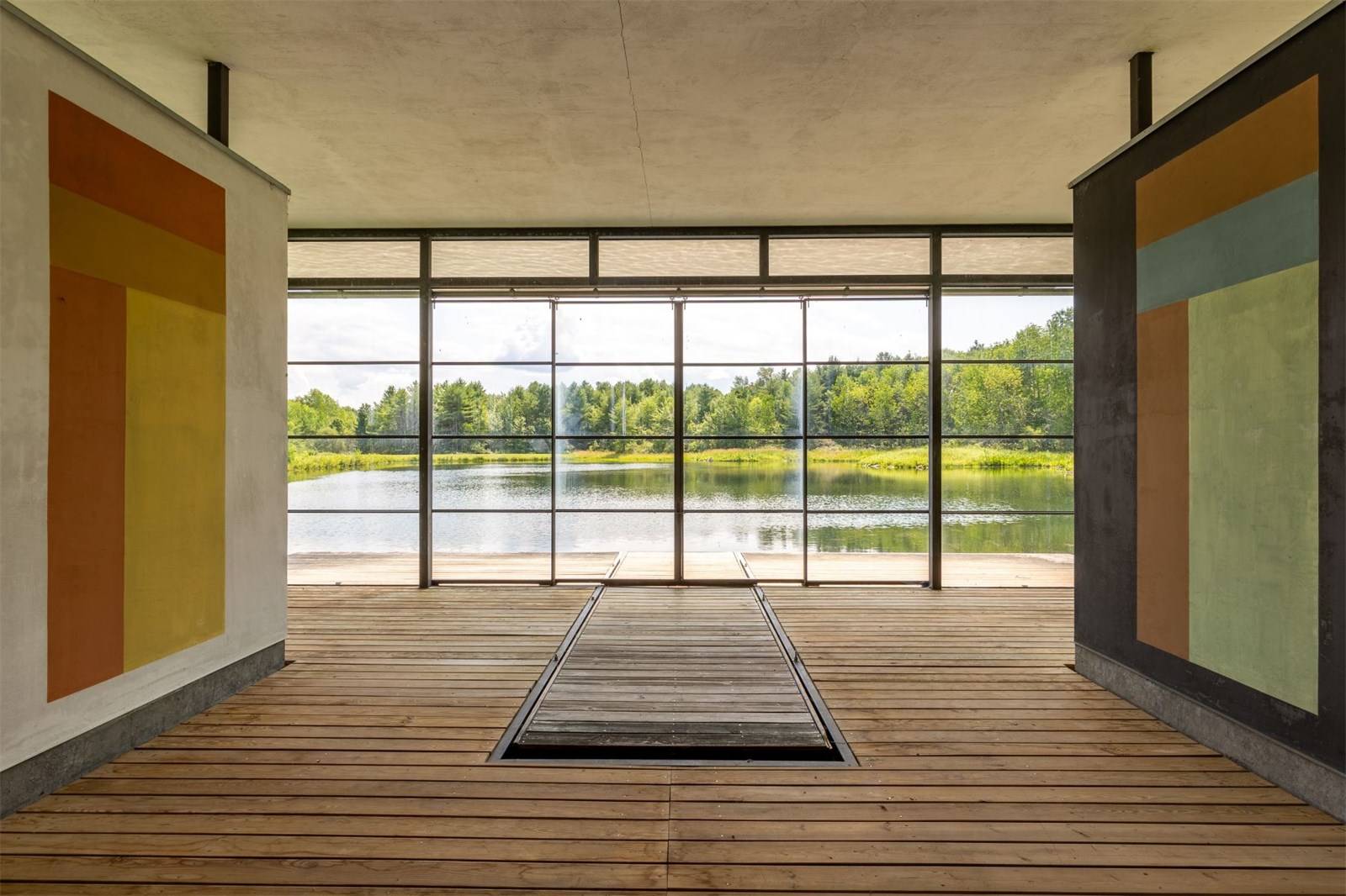 Room with large windows and colorful abstract panels, overlooking a serene lake and lush trees.