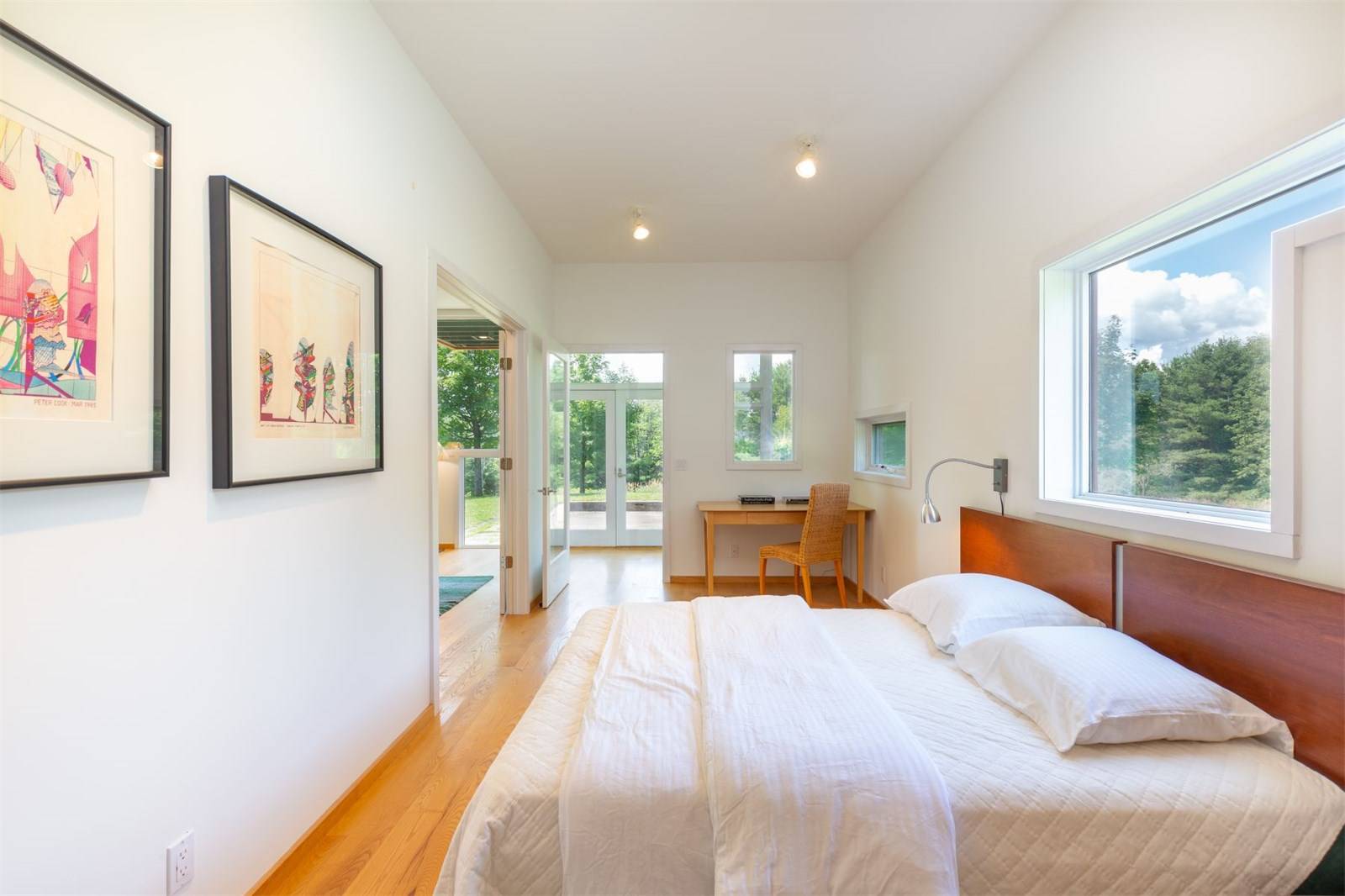 Minimalist bedroom with wooden floor, white bedding, wall art, desk, and large windows offering a view of greenery outside.