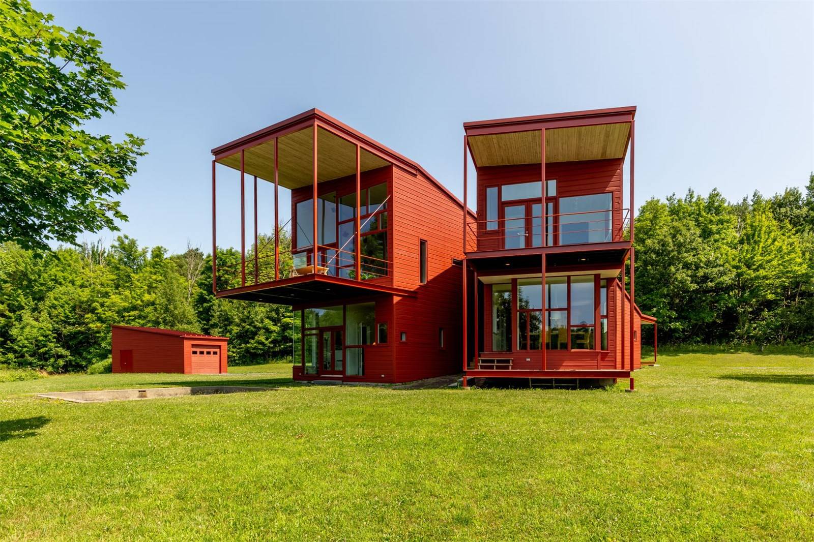 Modern red house with large windows on a green grassy landscape surrounded by lush trees under a clear blue sky.