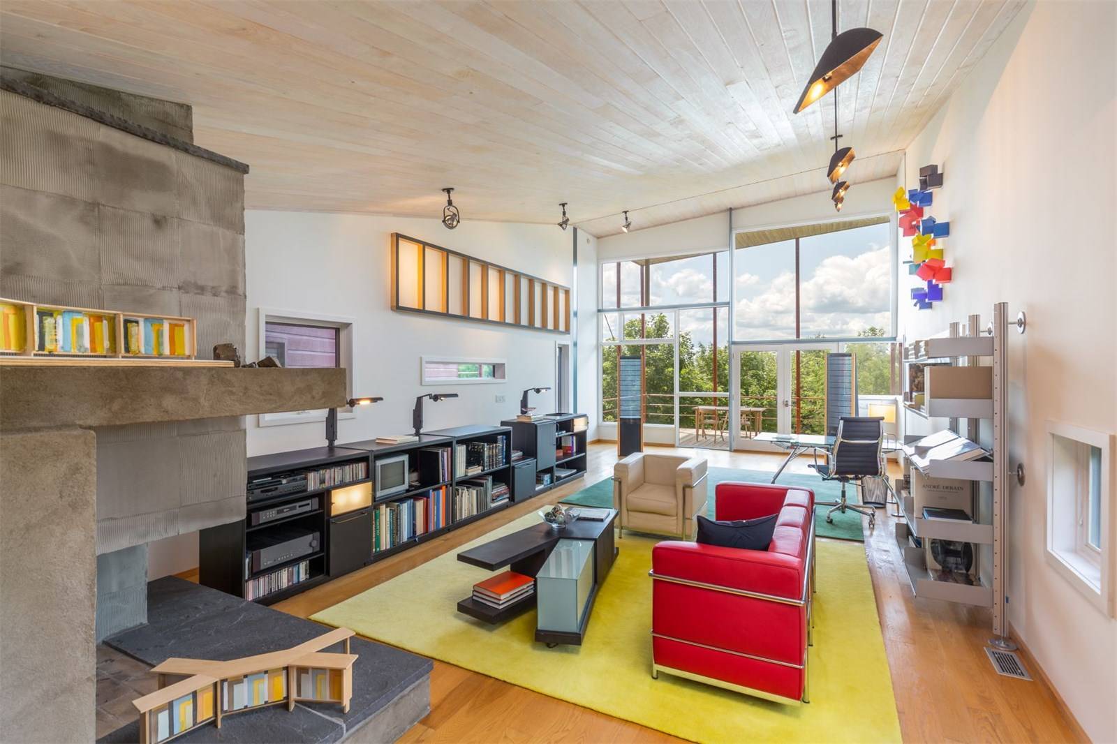 Modern living room with red sofa, large windows, bookshelves, and colorful wall art. Bright, open space with wooden ceiling.