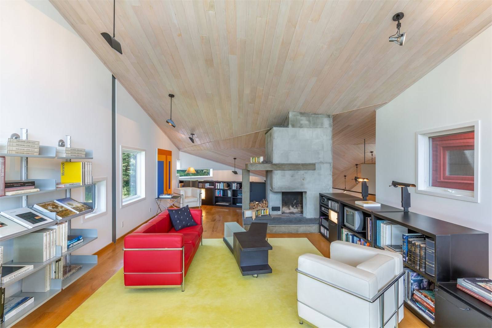 Modern living room with slanted wooden ceiling, red and white chairs, bookshelves, fireplace, and yellow rug.