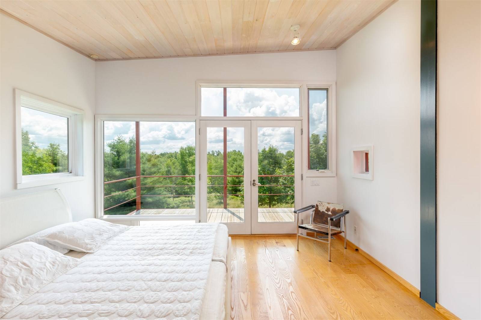 Bright and airy bedroom with wooden flooring, large bed, and views of trees through floor-to-ceiling windows and glass doors.