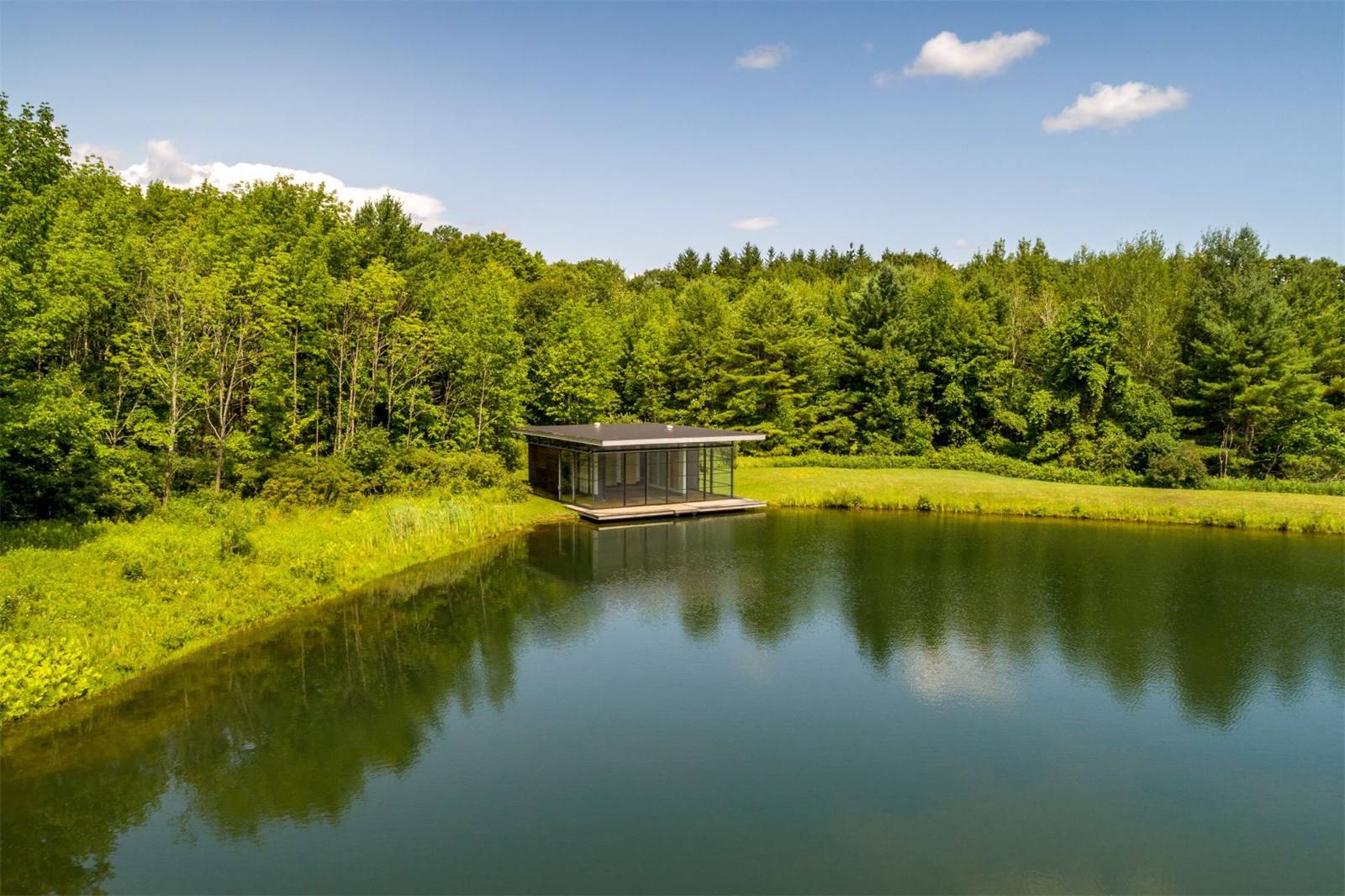 Modern glass-walled house by a tranquil lake surrounded by lush green trees under a clear blue sky.