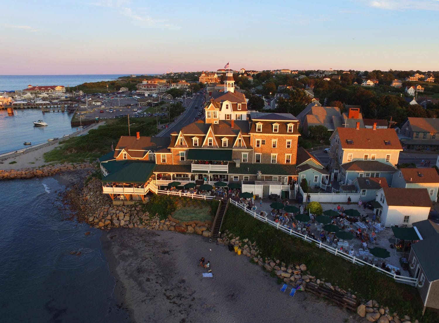 Aerial view of a seaside village with large building near a rocky beach and calm ocean at sunset.