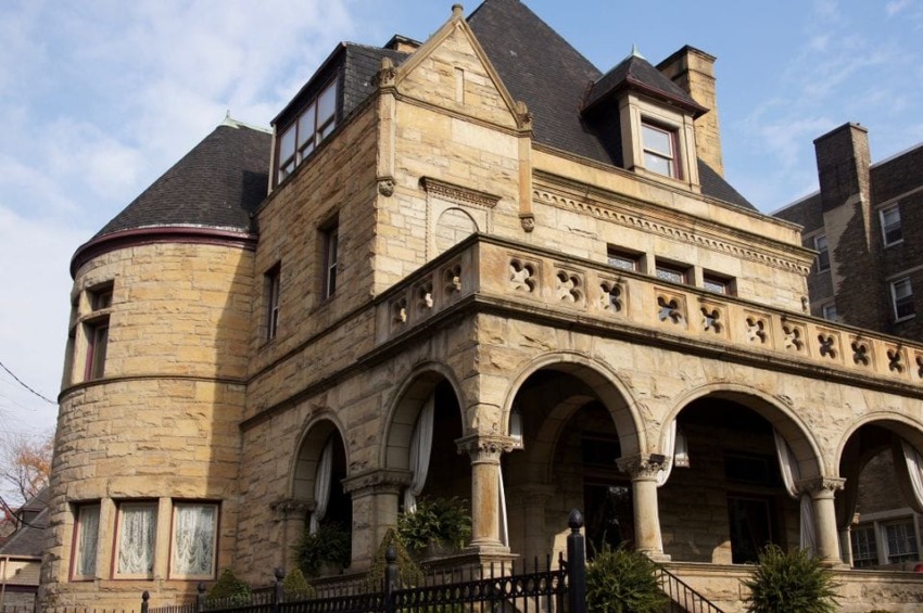 Historic stone mansion with arched porch, round tower, and intricate detailing under a clear blue sky.