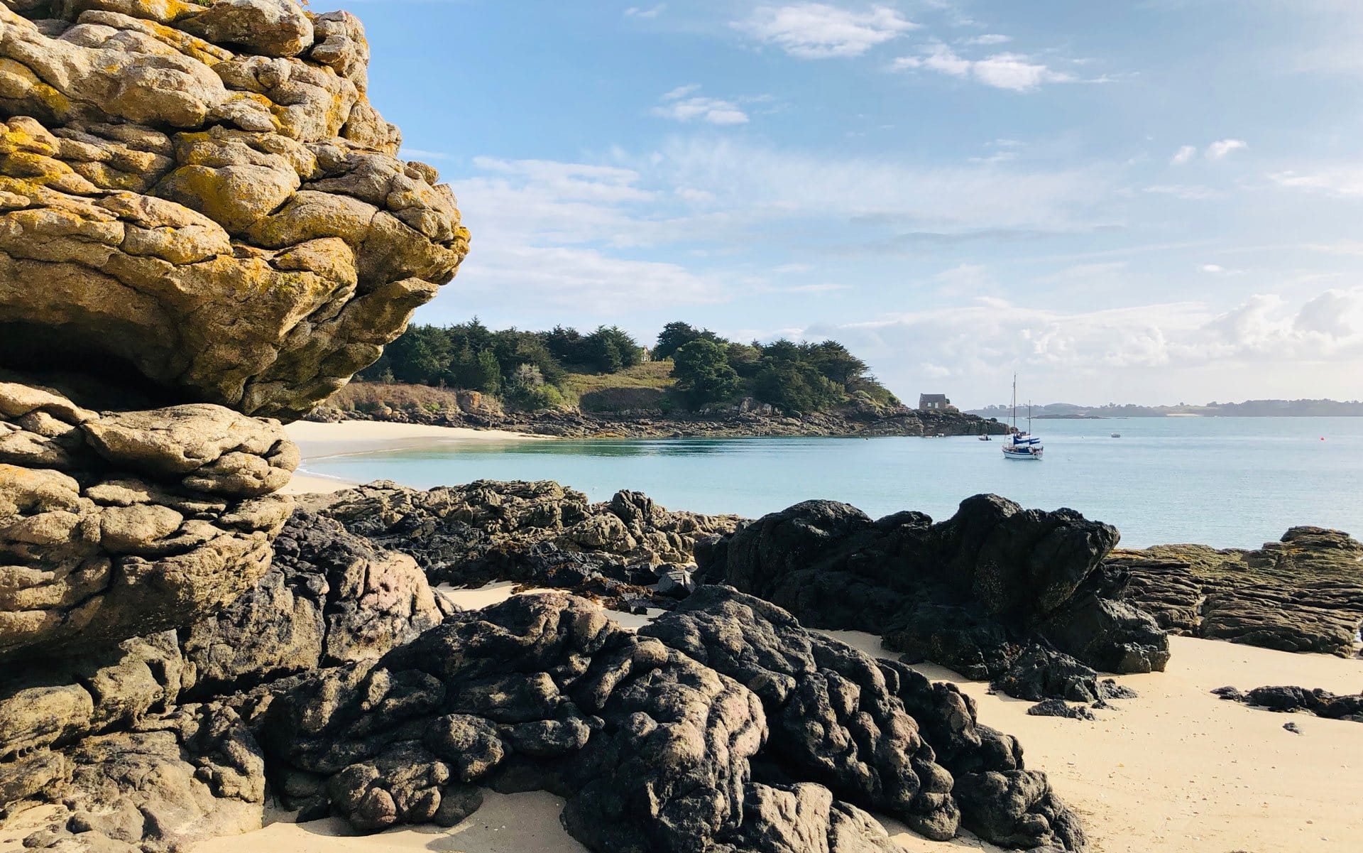 Rocky beach with clear water, distant sailboat, and lush greenery under a blue sky with scattered clouds.