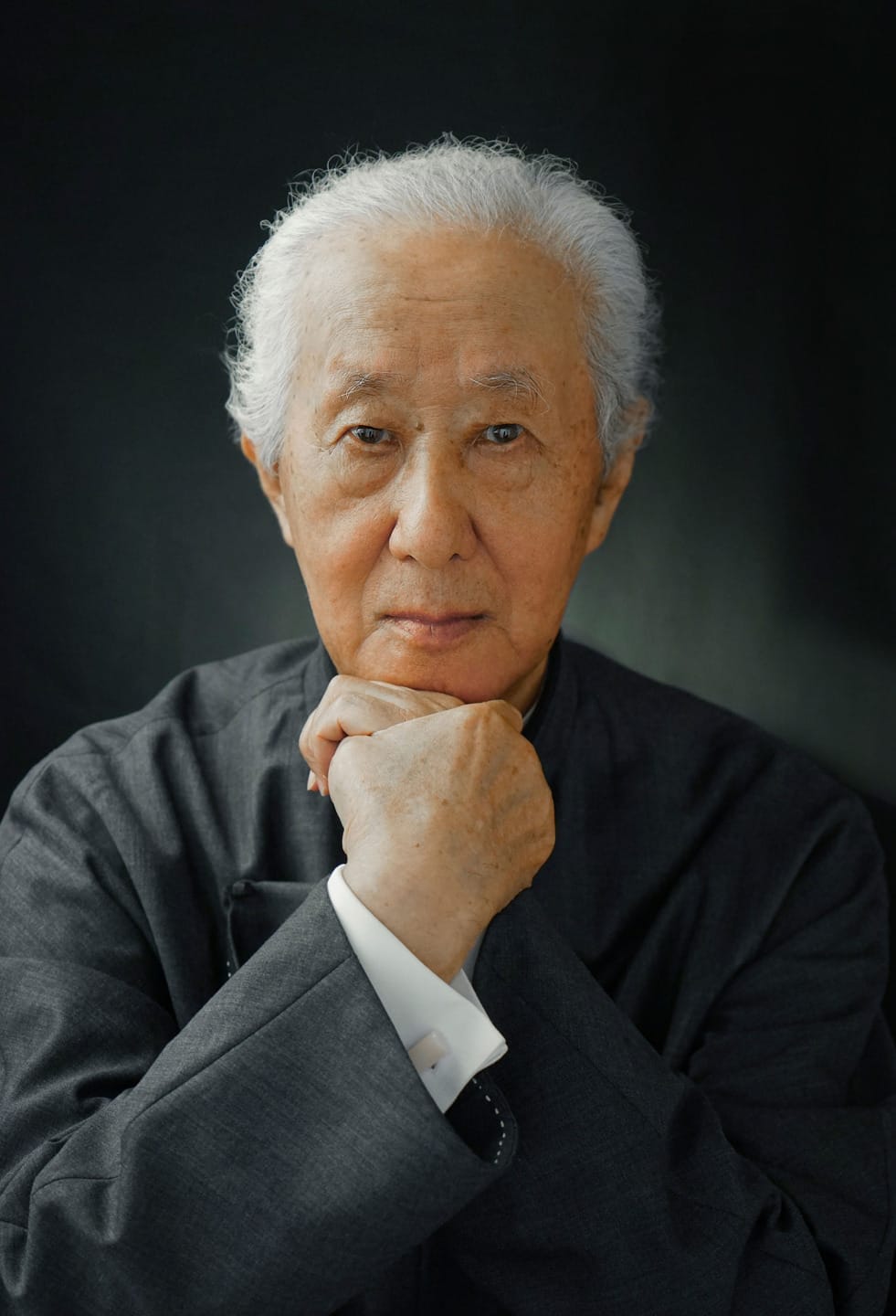 Elderly man with white hair wearing a dark suit, looking thoughtfully at the camera with hands under chin against a dark background.