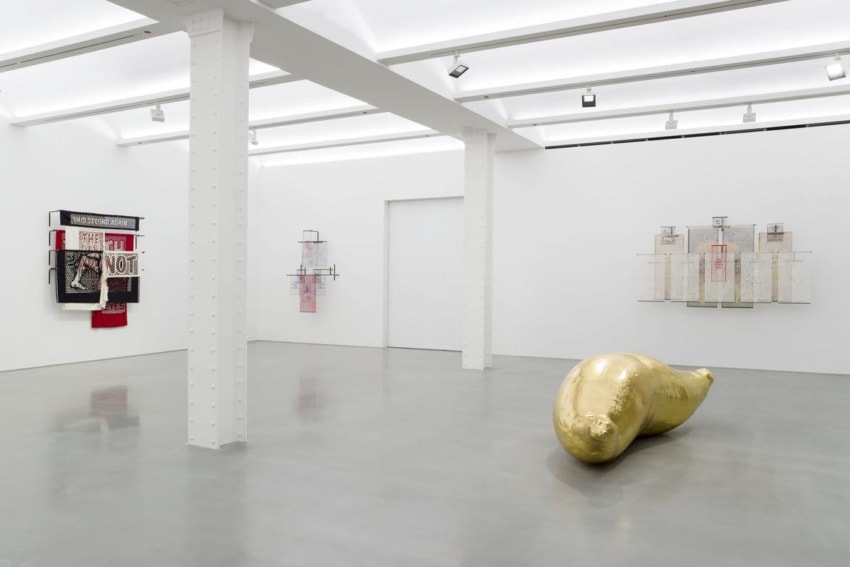 Contemporary art gallery with sculptures, mixed media artworks on white walls, and a large golden sculpture on the floor.