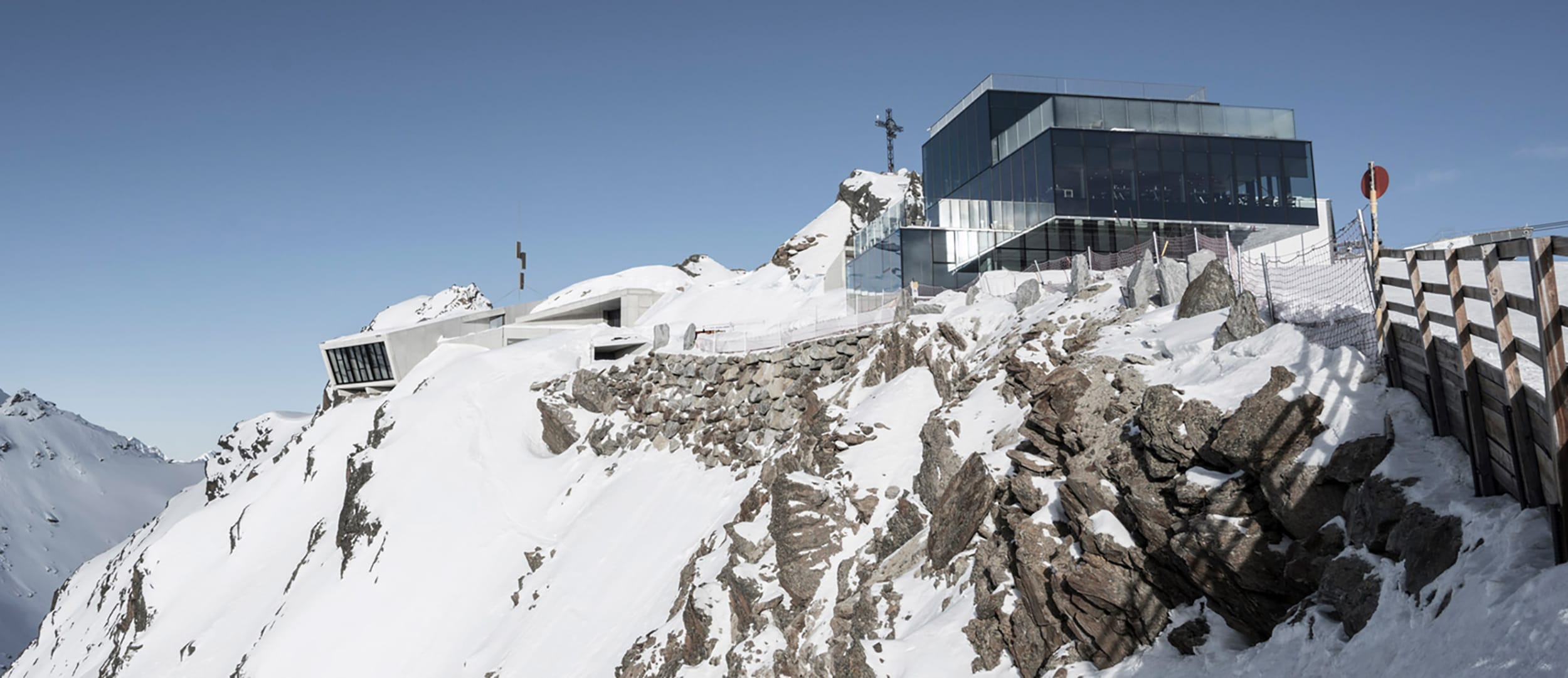 Modern glass building perched atop a snowy mountain peak with a clear blue sky in the background.