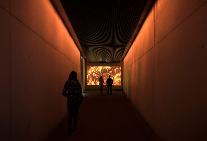 People walking in a dimly lit tunnel toward a large bright screen displaying fiery abstract art at the end.