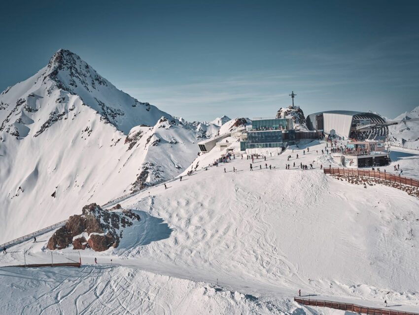 Ski resort on a snowy mountain landscape with a clear blue sky and people skiing near a modern building complex.