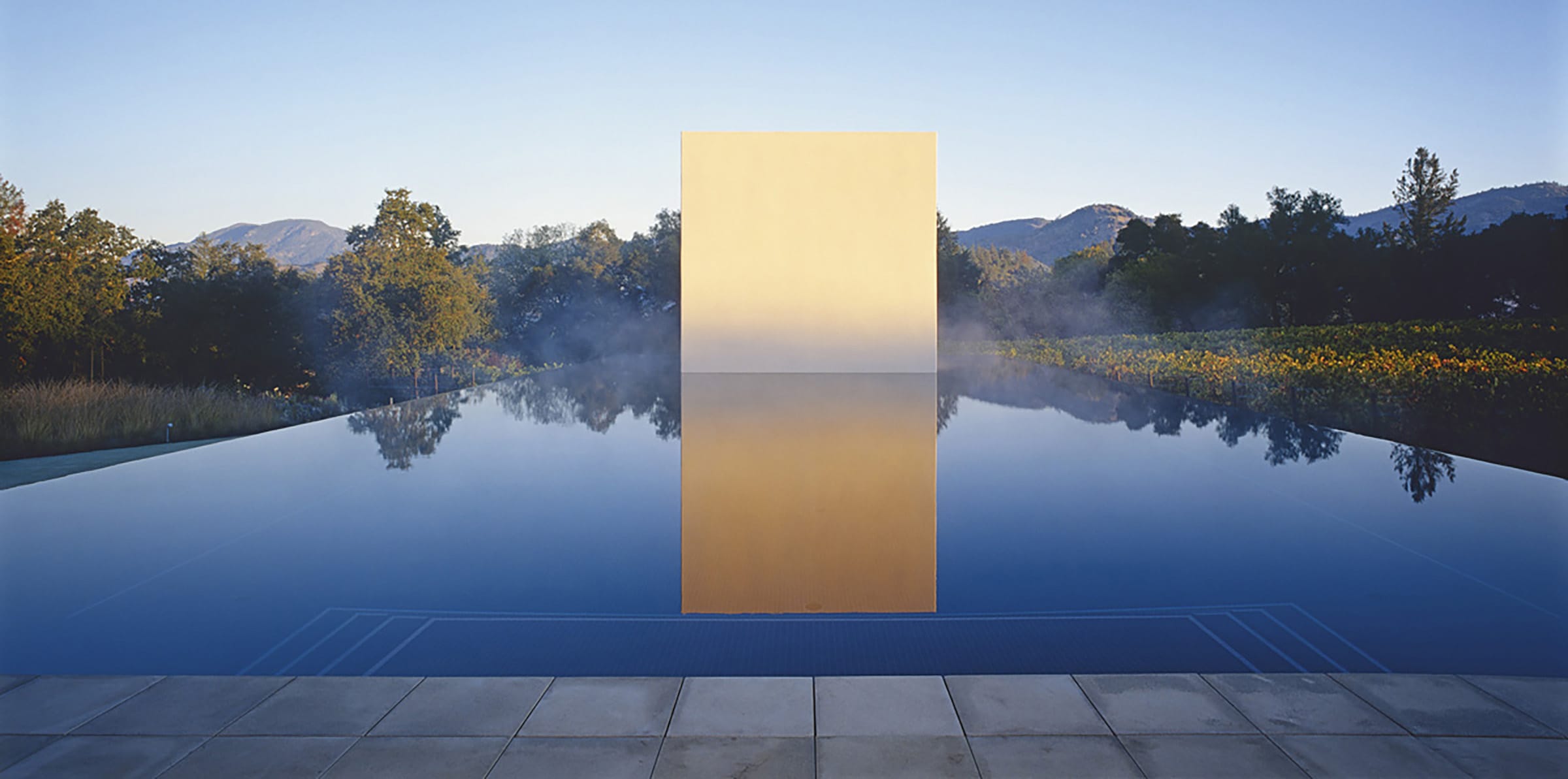 Reflective pool with large golden geometric structure in center, surrounded by mist, trees, and mountains in the background.