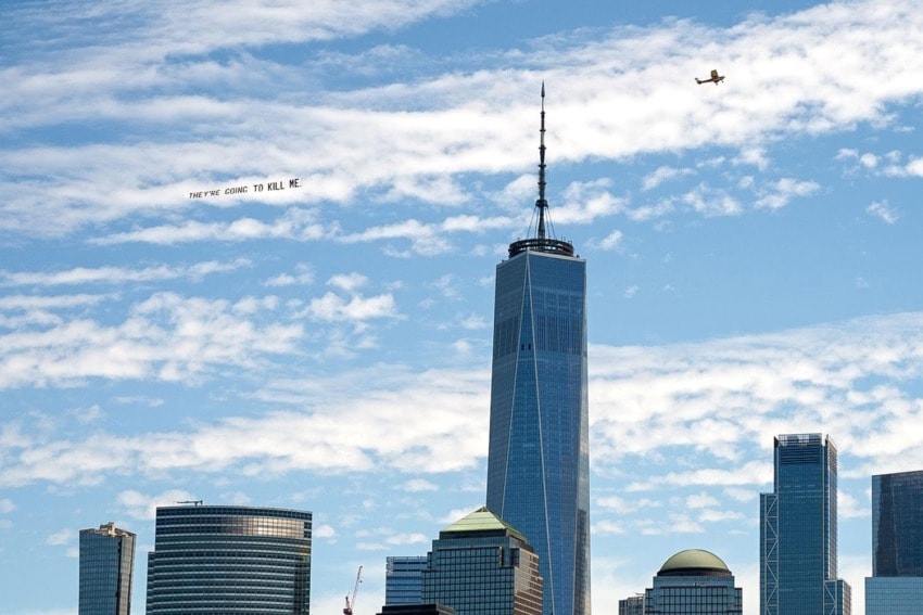 Plane flying over New York City skyline with a banner reading a distressing message, One World Trade Center visible.