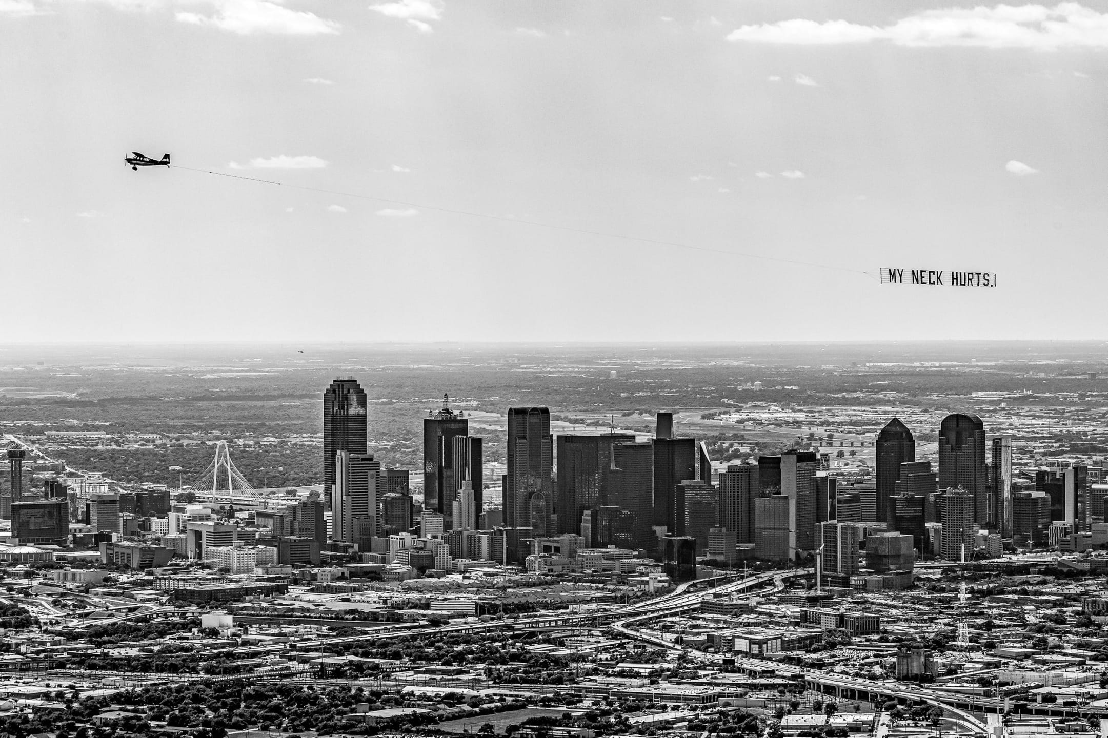 Black-and-white cityscape with an airplane towing a banner that reads, "MY NECK HURTS," over the skyline.