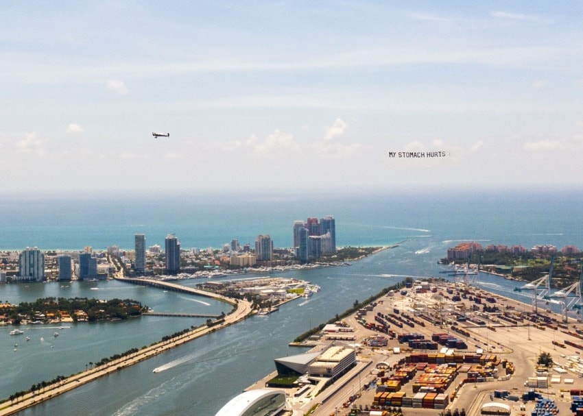 Aerial view of a city by the sea with "MY STOMACH HURTS" written in the sky and a passing airplane.