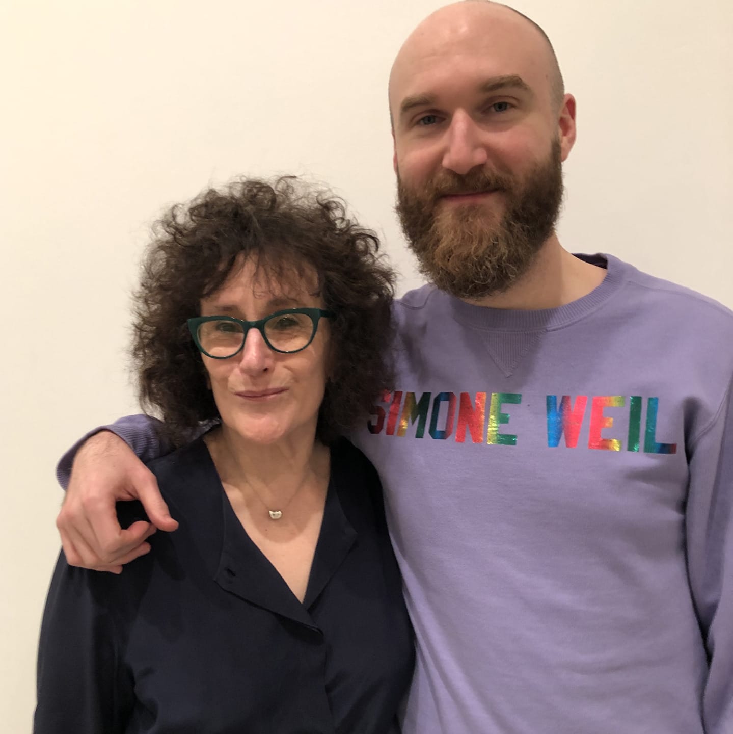 A woman with curly hair wearing glasses and a man with a beard in a colorful shirt, posing together in front of a white wall.