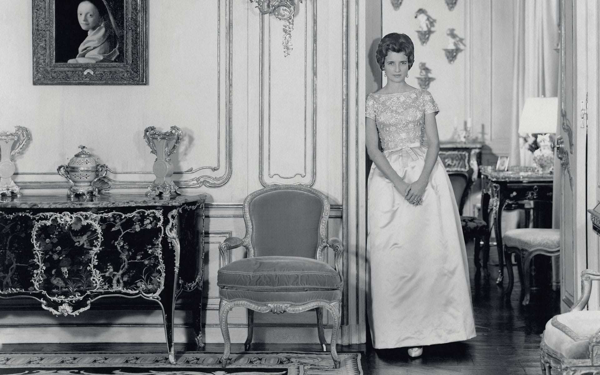 Woman in an elegant dress standing in an ornate room with classic furniture and a portrait on the wall.