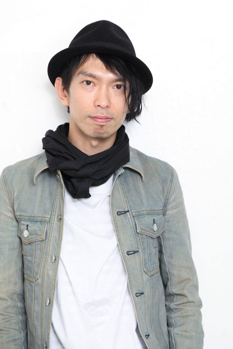 Person wearing a denim jacket, white shirt, black scarf, and black hat, standing against a white background.