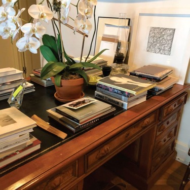 Wooden desk with stacks of books, a potted orchid, and framed artwork in a well-lit room.