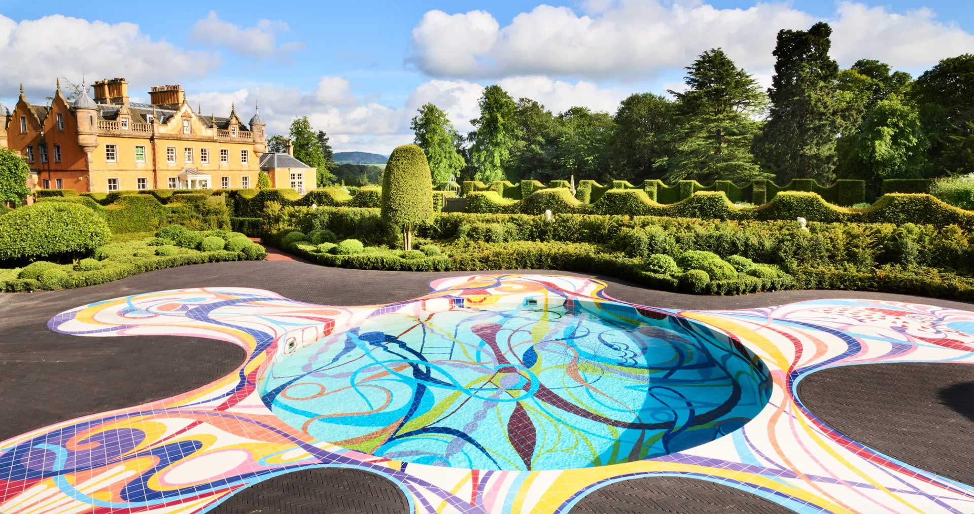 Colorful mosaic water feature in ornate garden with historic building and lush greenery in background under a blue sky.