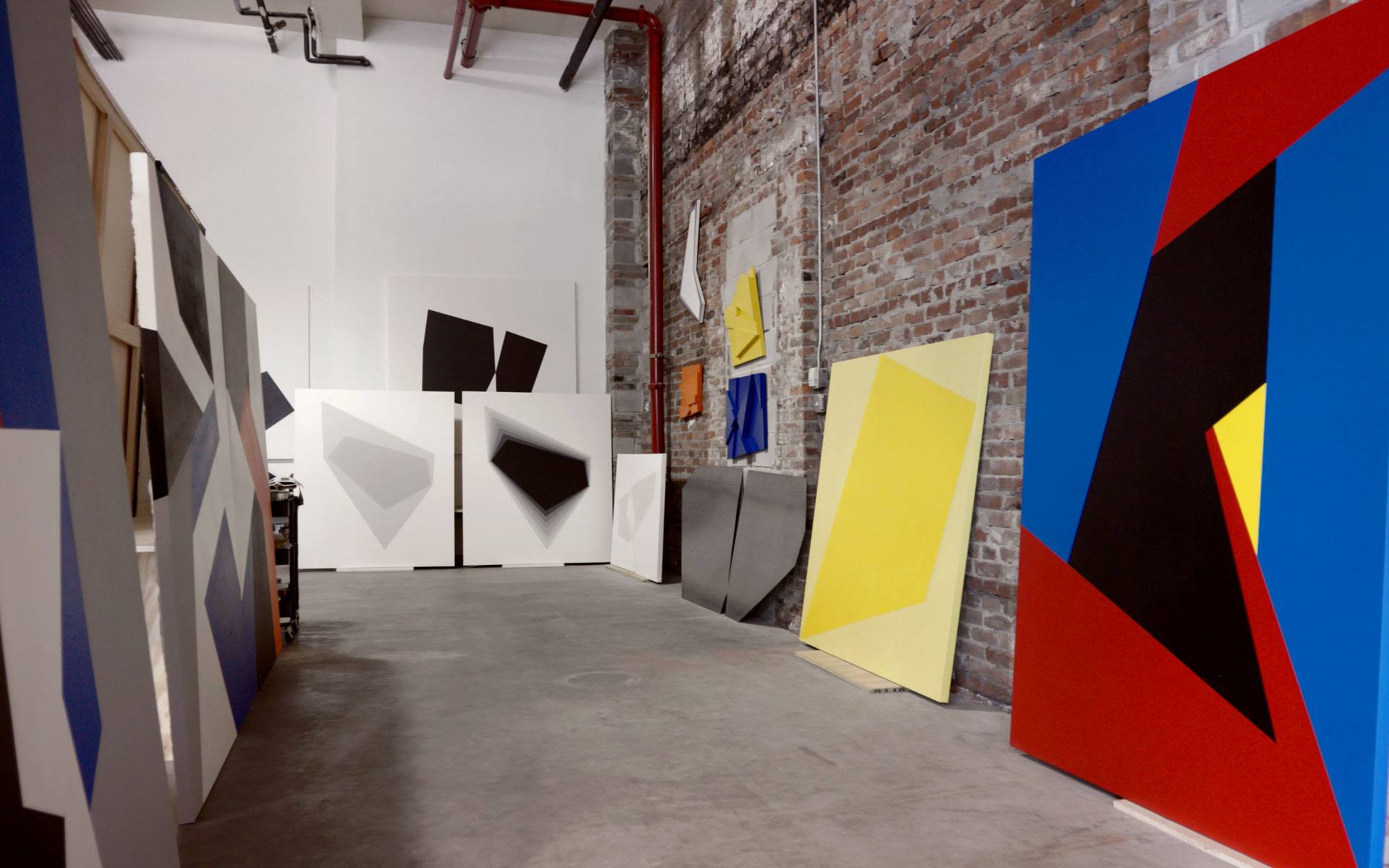 Art studio with abstract geometric paintings displayed on large canvases against brick wall and floor.