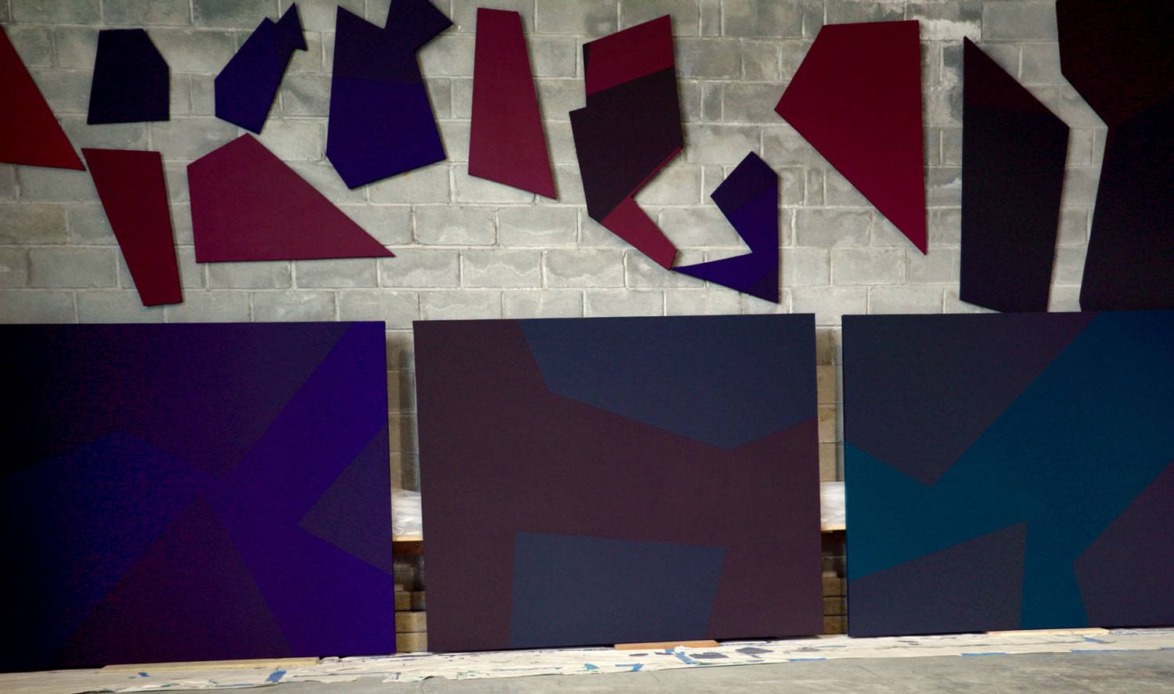 Abstract geometric art pieces on a concrete wall, featuring dark shades of purple, maroon, and blue shapes.