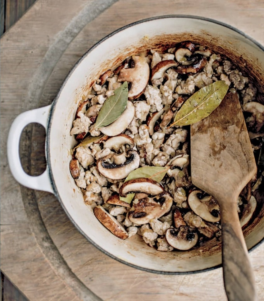 Rustic pot with cooked sausage, mushrooms, and bay leaves on a wooden table, stirred with a wooden spatula.