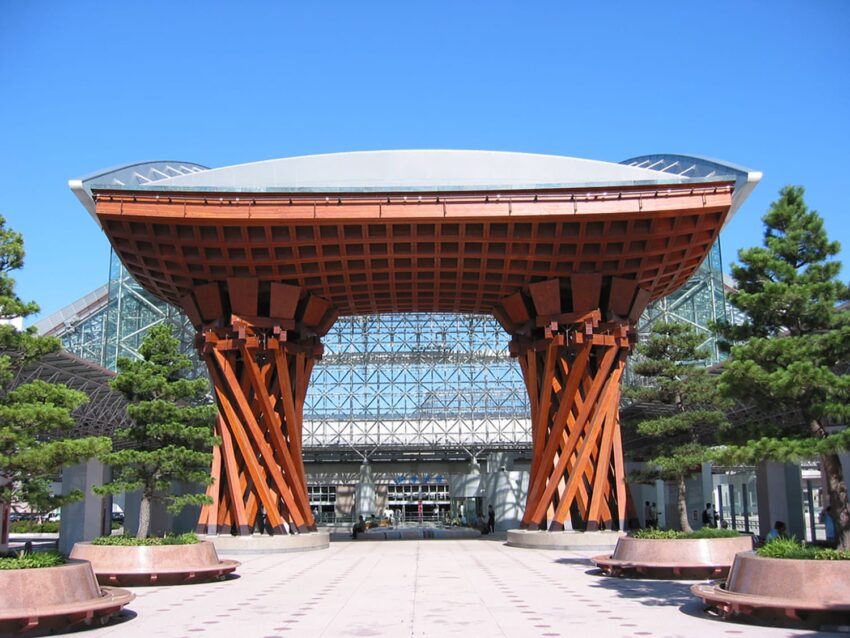 Large wooden gate structure with lattice design at entrance of modern building, surrounded by trees and clear blue sky.