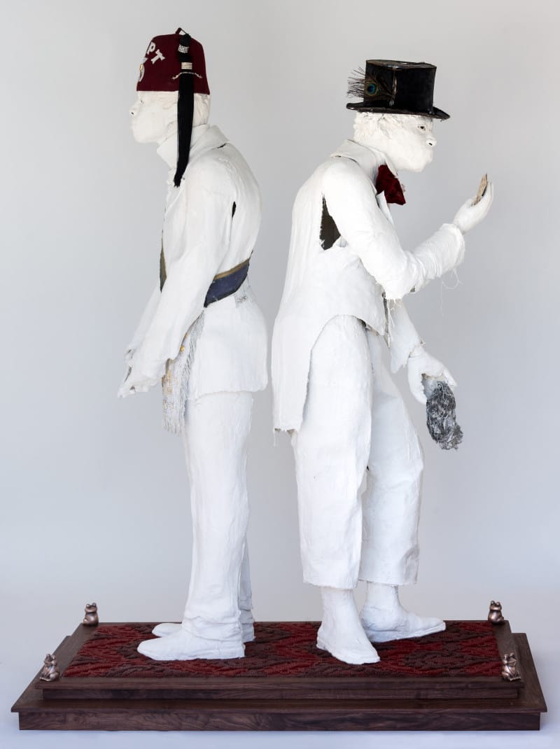 Two life-sized, white sculptures wearing hats, standing back-to-back on a decorative platform with a red pattern.