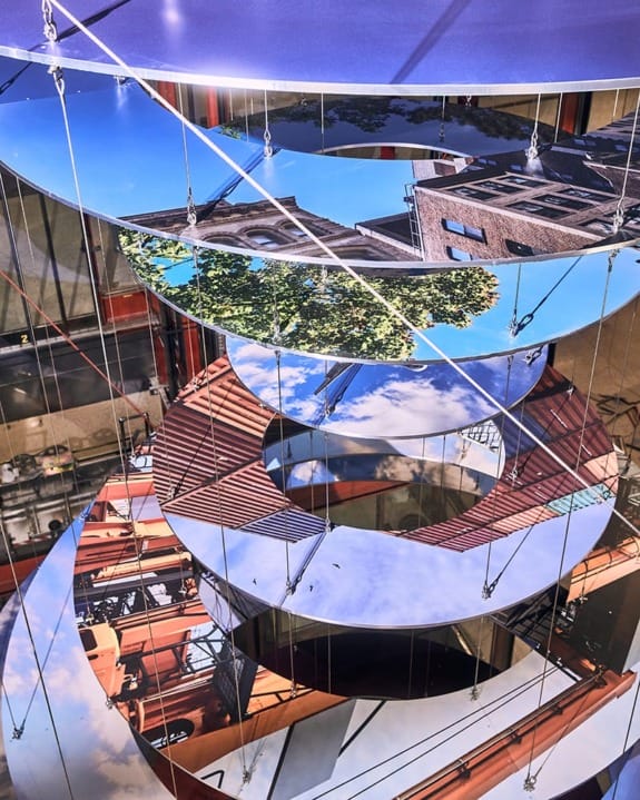 Artistic installation of suspended circular mirrors reflecting urban scenes and blue sky in vertical spiral arrangement