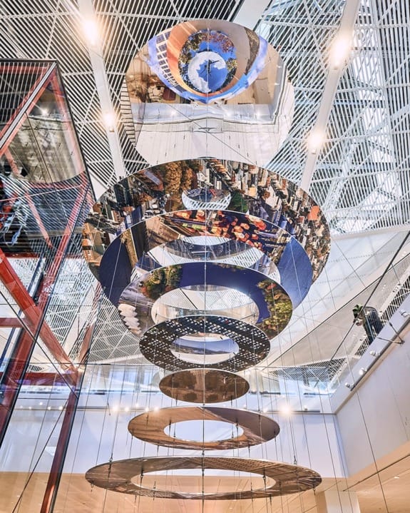 Suspended art installation with circular mirrors reflecting cityscapes and ceiling architecture in a modern building interior.