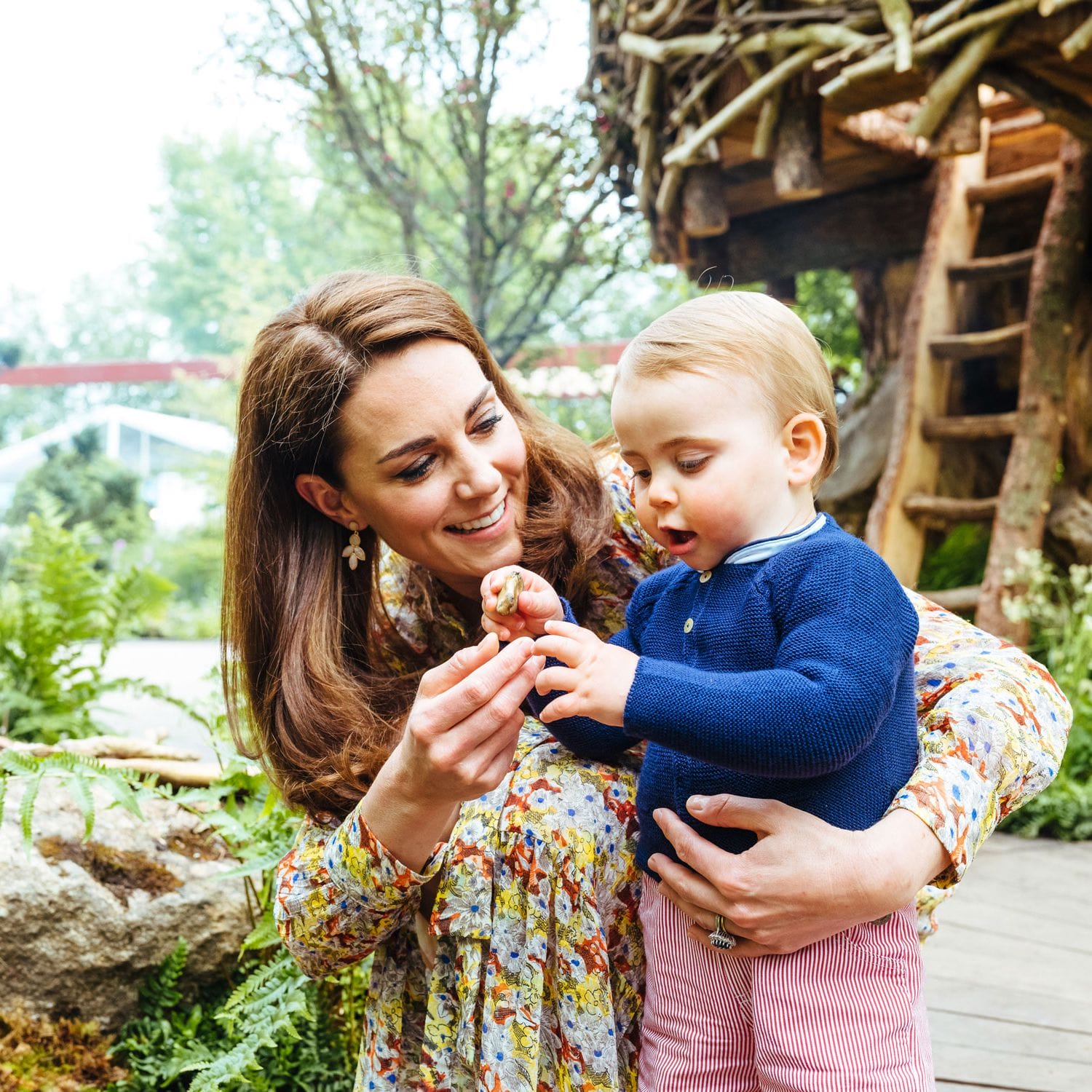 Woman in floral dress and a young child examine a small object outdoors near a wooden structure and greenery.