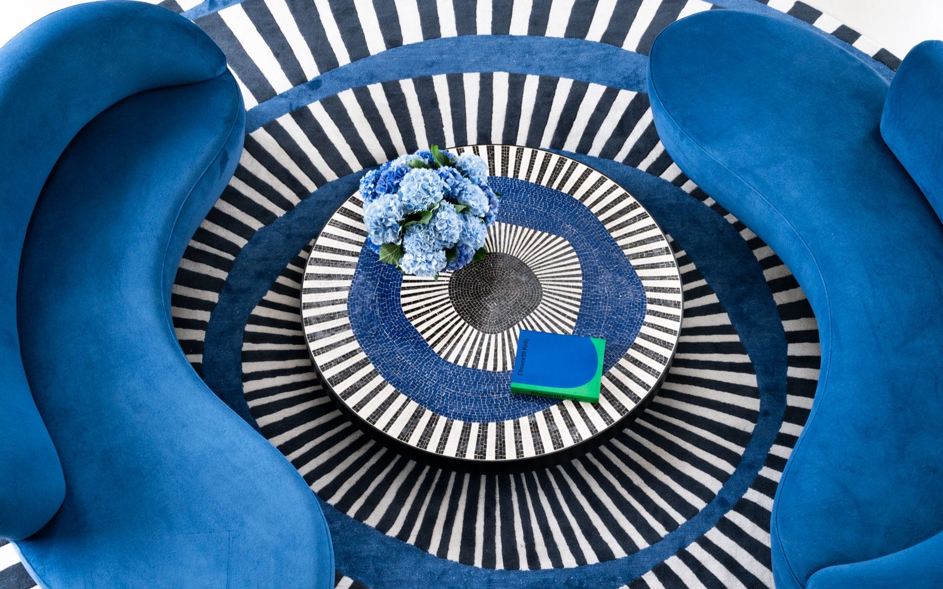 Modern living room with blue velvet sofas, circular striped rug, round table, and blue hydrangea centerpiece.