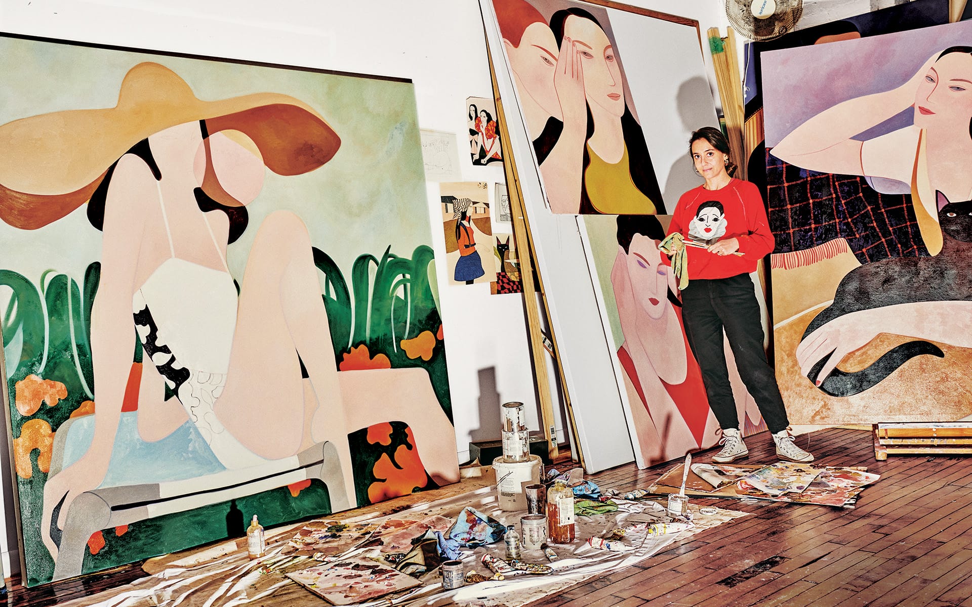 Artist standing in a studio with large colorful paintings, surrounded by art supplies on the floor.