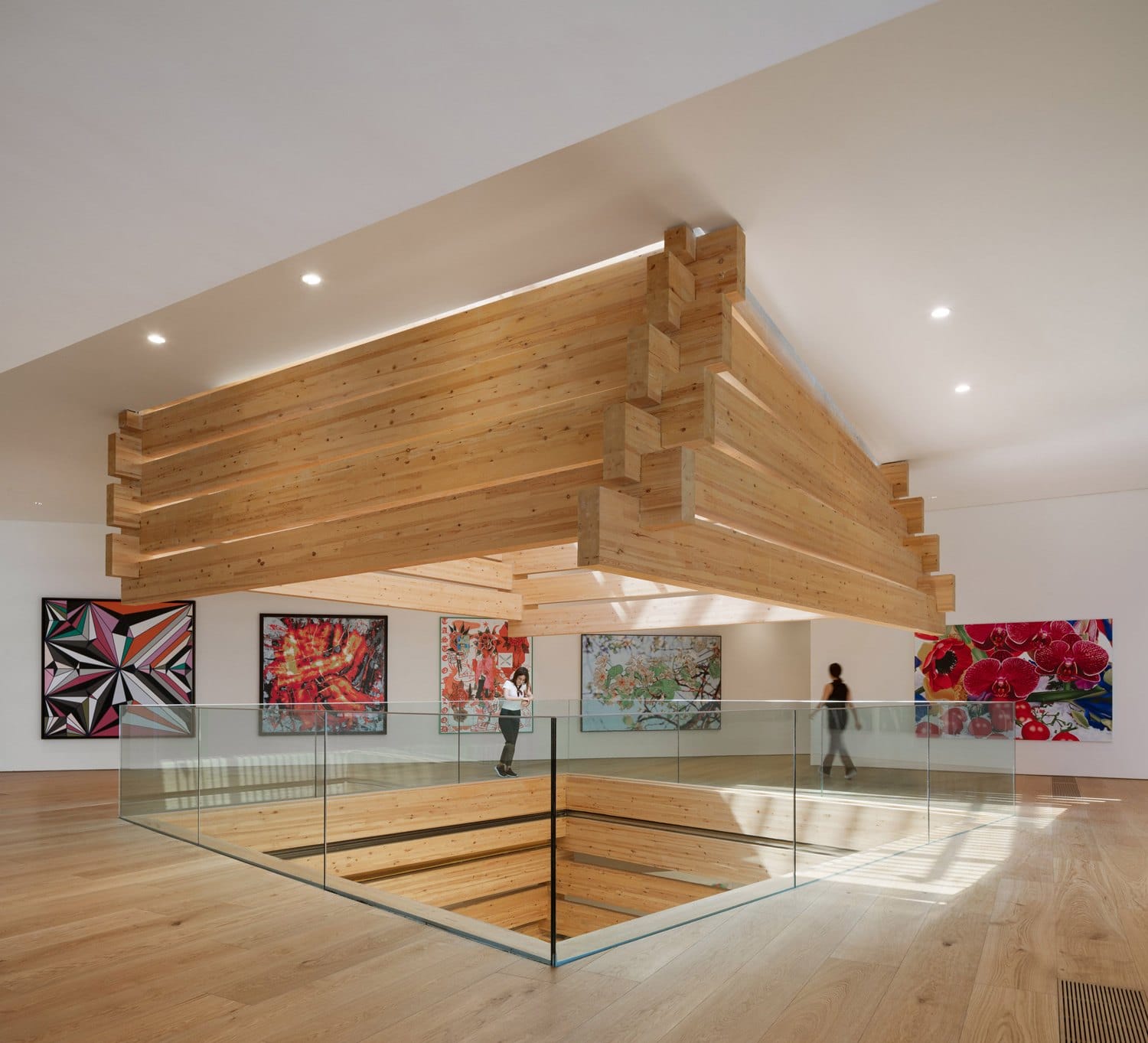 Modern art gallery interior with abstract paintings, large wooden ceiling centerpiece, and visitors observing the exhibits.
