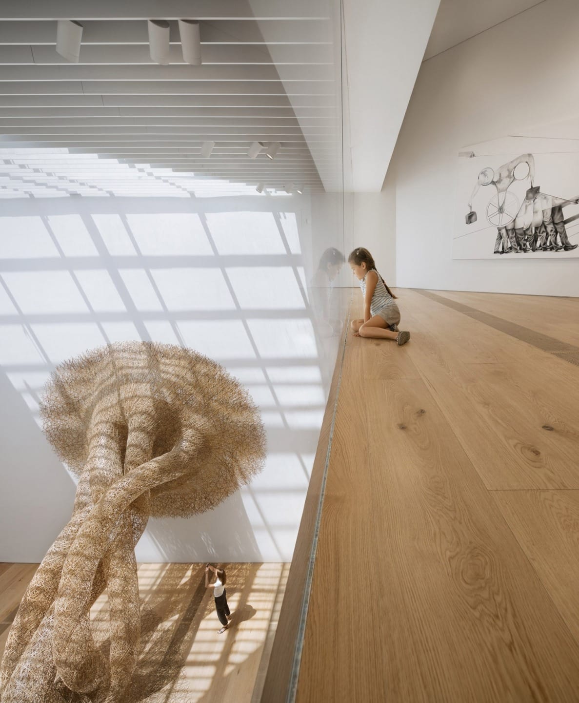 Child observing large sculptural installation in a modern art gallery with wooden floors and high ceilings, sunlight casting shadows.