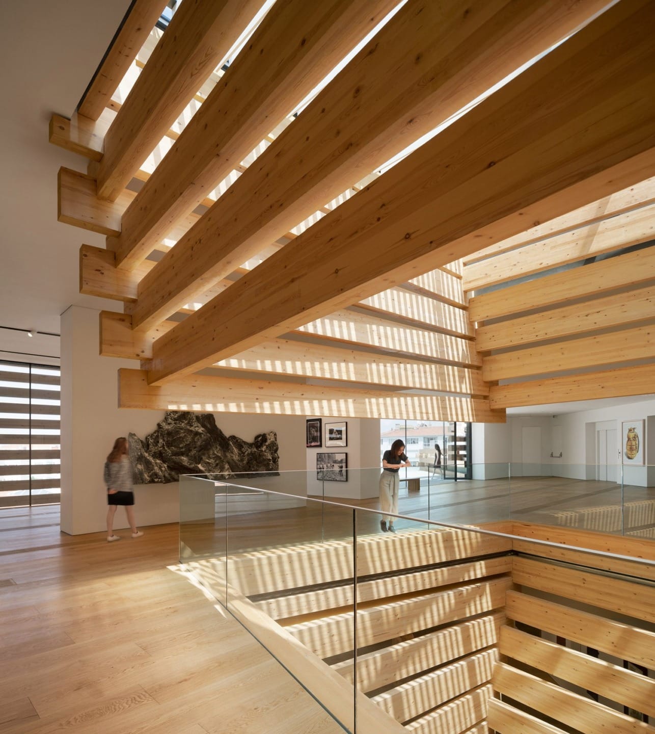 Modern interior with wooden beams, glass railings, and natural light streaming in; visitors walking and observing artwork.