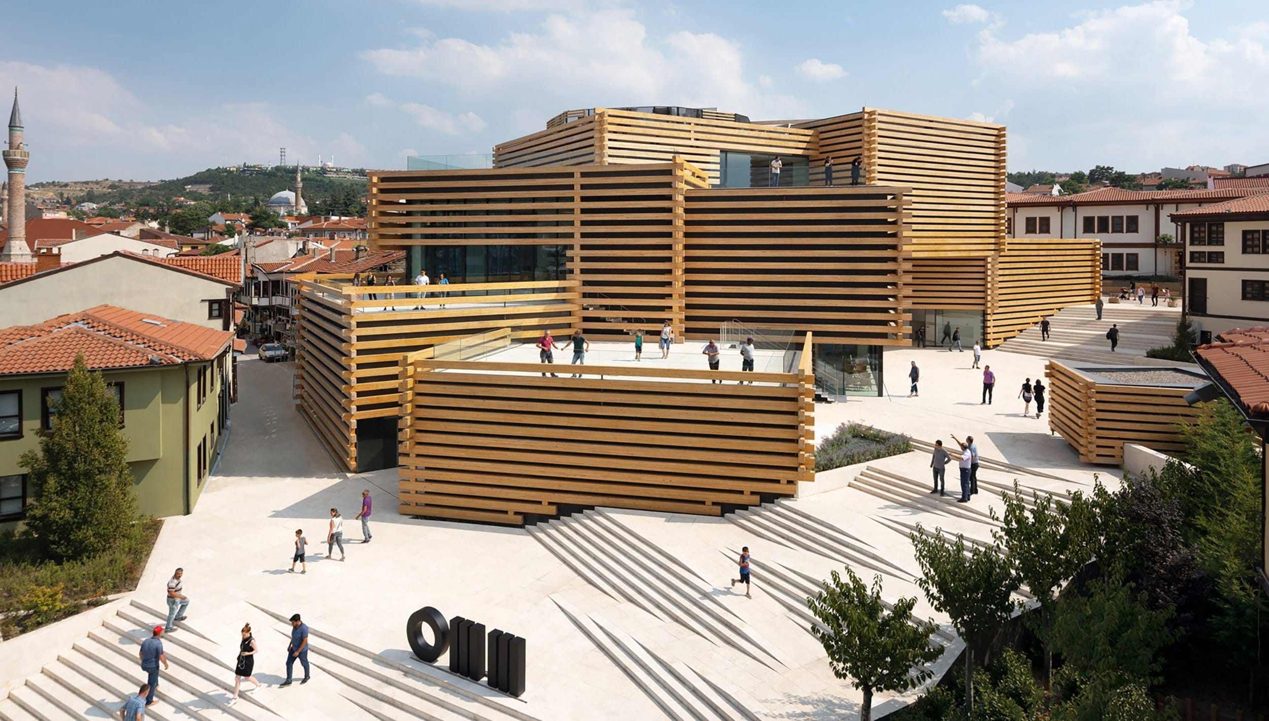 Modern multi-level building with horizontal wooden panels, surrounded by people on stairs, located in a sunny urban area.
