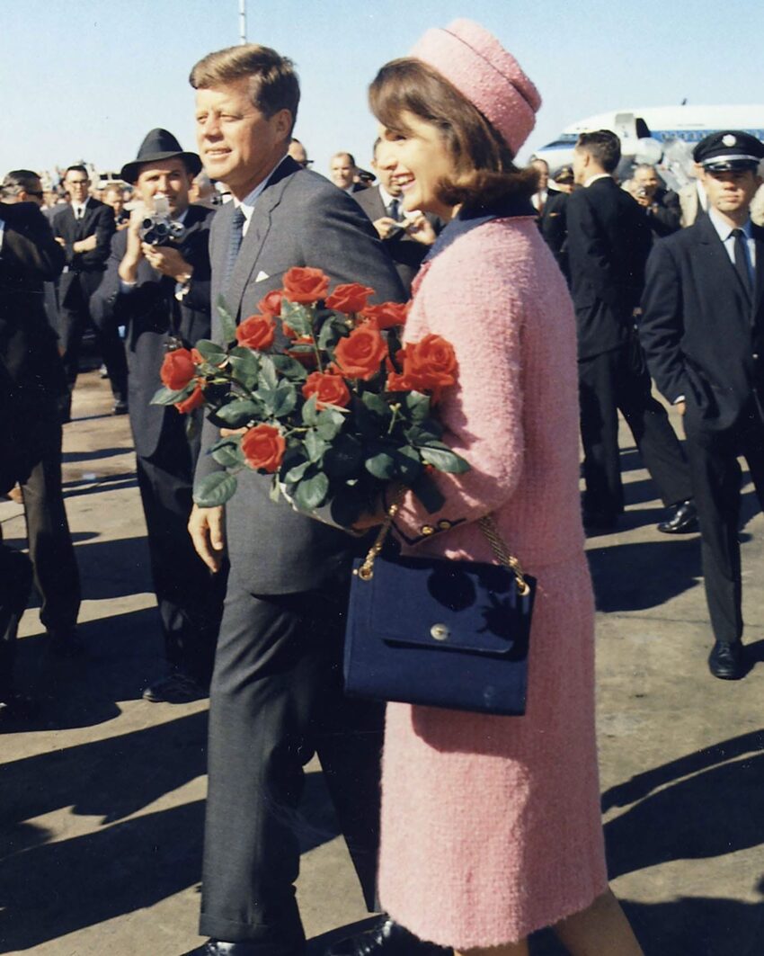 A woman in a pink outfit holding a bouquet of red roses standing next to a man in a suit amidst a crowd.