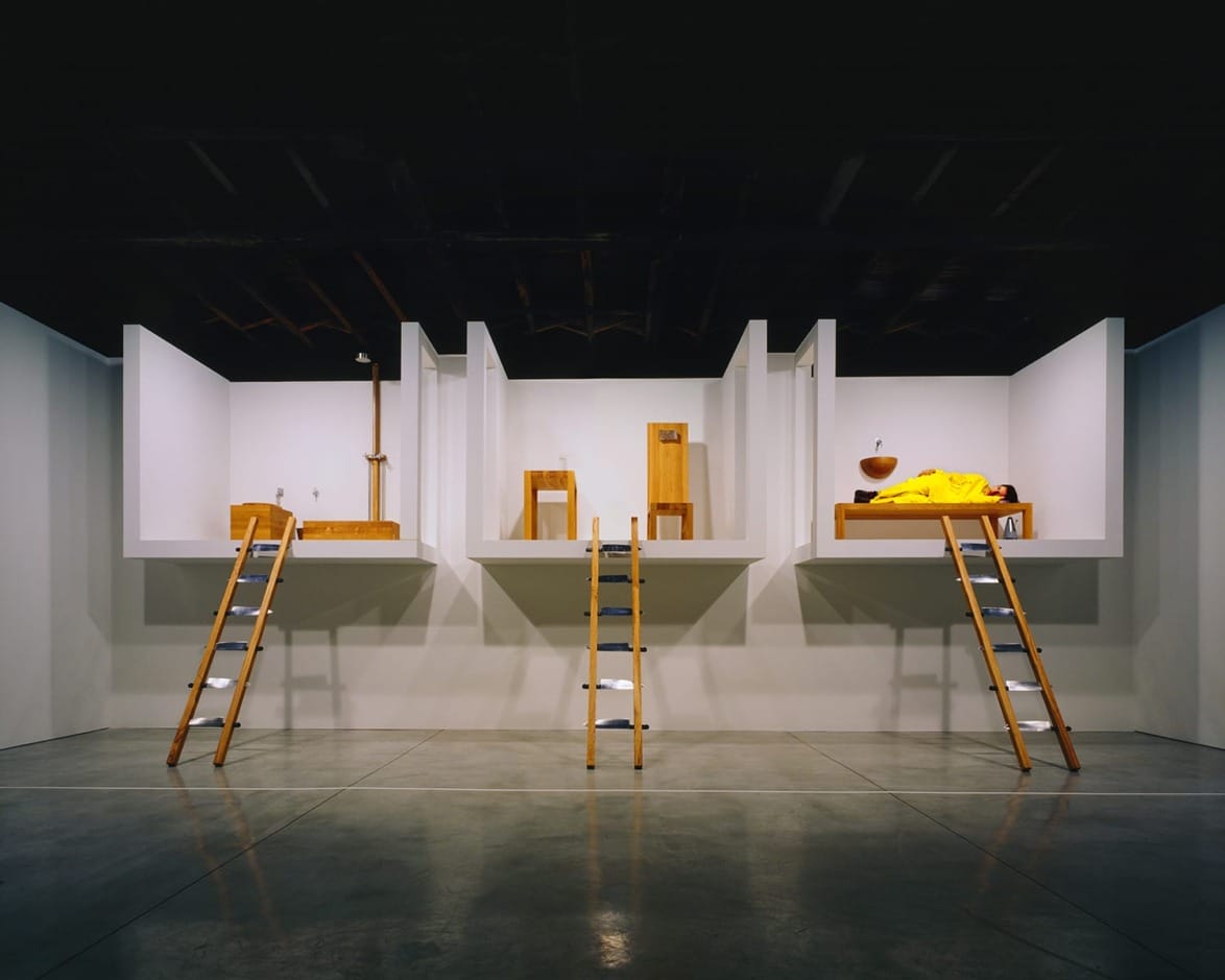 Art installation with three wooden ladders leading to minimalistic rooms, each featuring a bed, chair, and table.