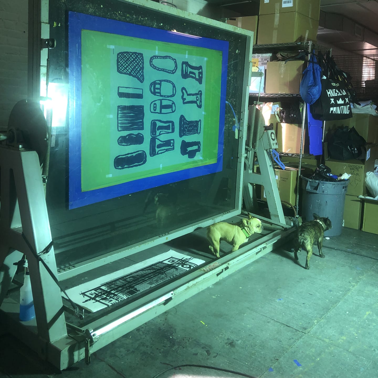 Two dogs exploring a large screen printing setup in a cluttered workshop with various supplies and equipment.