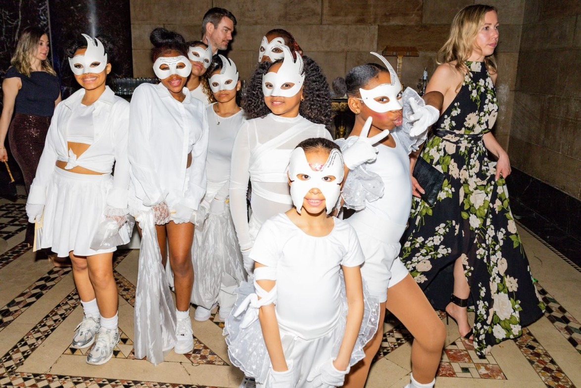 Group of people in white costumes and masks, posing on a patterned floor with a woman in a floral dress nearby