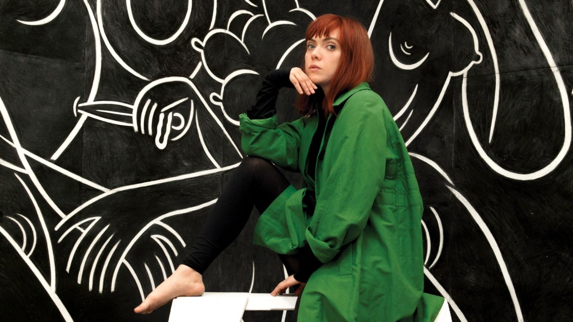 Person in green coat seated in front of a black and white abstract mural.
